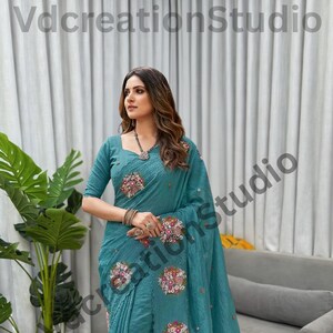 May include: Teal saree with floral embroidery. The saree features intricate floral designs in shades of pink, white, and red. The sleeves are three-quarter length. The text "VdcreationStudio" is visible in the image.