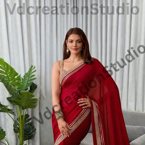 Peut inclure: Un sari rouge foncé avec des bordures dorées et un chemisier assorti. Le sari est drapé avec élégance, mettant en valeur un design fluide. Le texte "VdcreationStudio" est inclus.