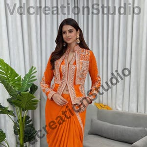 May include: An orange saree and matching jacket with intricate embroidery. The jacket has a high collar and long sleeves. The saree is draped elegantly, and the outfit is accessorized with gold earrings. The outfit is on a round rug.