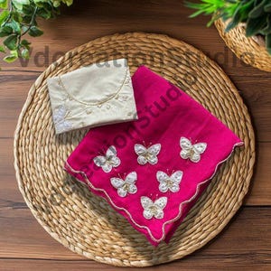 May include: A vibrant fuchsia handkerchief, adorned with embroidered white butterflies and gold edging, alongside a cream-coloured pouch. Both items are displayed on a woven mat. The butterfly motif is a key design element.