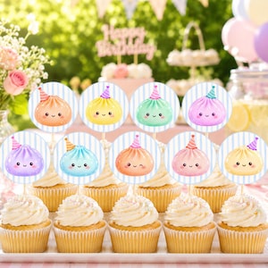 May include: A set of nine cupcakes arranged on a white rectangular tray, each topped with a circular paper topper featuring a colorful cartoon dumpling character wearing a party hat against a light blue and white vertical striped background.