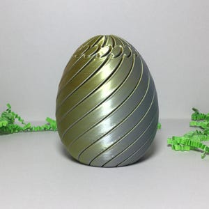 May include: A decorative egg-shaped object with a metallic gradient finish, transitioning from gold to silver. The egg features a series of diagonal, curved lines. Green paper grass is scattered around the base of the egg.
