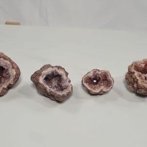 May include: Four amethyst geodes with a rough, textured exterior and a crystalline interior. The geodes vary in size, with the largest measuring approximately 4 inches across. The color palette includes shades of purple, brown, and beige.