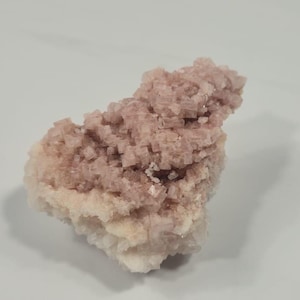 Puede incluir: Primer plano de un espécimen mineral con un grupo de cristales rosas y blancos. Los cristales tienen forma cúbica, con un tono rosa pálido en la parte superior y una base blanca. El fondo es blanco.