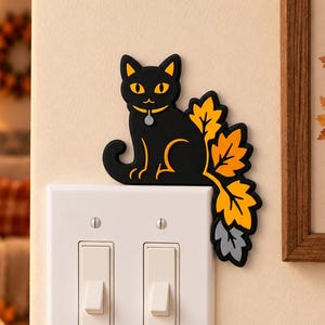 以下が含まれることがあります： 黄色い目と小さな灰色の首輪タグが付いた黒猫のウォールステッカー。白いダブルライトスイッチプレートの上に配置されています。猫の尻尾は右側に向かってオレンジ、黄色、灰色の秋の葉の集まりへと続いています。