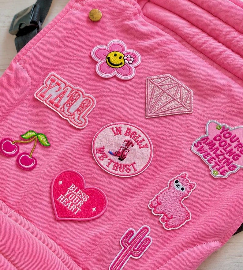 K&ouml;nnte beinhalten: Ein pinkfarbener Stoffartikel mit verschiedenen gestickten Aufn&auml;hern. Zu den Aufn&auml;hern geh&ouml;ren eine Blume mit einem Smiley, ein Diamant, ein Herz mit "Bless Your Heart" und ein Kaktus. Andere Aufn&auml;her zeigen Text und ein rosa Lama.