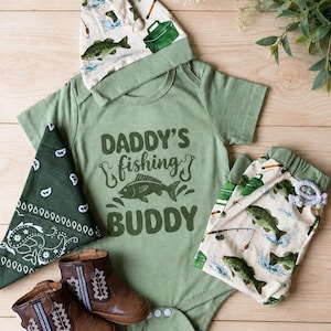 May include: A baby outfit set in shades of green and brown, featuring a bodysuit with "Daddy's Fishing Buddy" text and a fish graphic. The set includes a matching hat, pants, bandana, and brown leather boots. The hat and pants have a fish pattern.