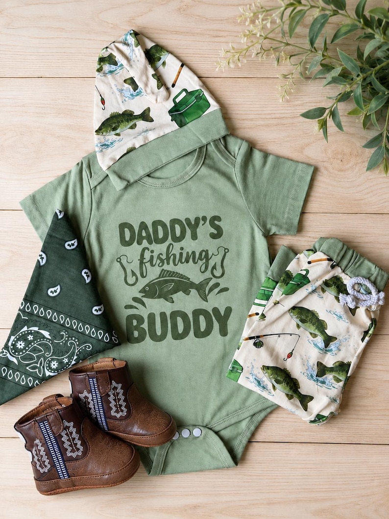 May include: A baby outfit set in shades of green and brown, featuring a bodysuit with "Daddy's Fishing Buddy" text and a fish graphic. The set includes a matching hat, pants, bandana, and brown leather boots. The hat and pants have a fish pattern.