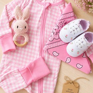 May include: A baby clothing set in pink and white gingham includes a zippered onesie, a bandana, and a pair of white shoes with floral embroidery. A crochet bunny rattle and a tag that says "made with love" complete the set.