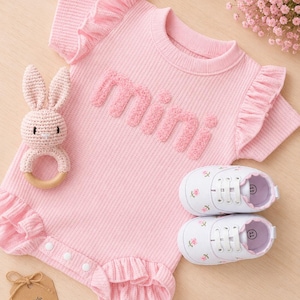 K&ouml;nnte beinhalten: Ein rosa Baby-Strampler mit R&uuml;schen&auml;rmeln und dem Wort "mini" in strukturierten Buchstaben. Enth&auml;lt eine H&auml;schen-Rassel, wei&szlig;e Babyschuhe mit Blumenstickerei und ein Etikett mit der Aufschrift "made with love".