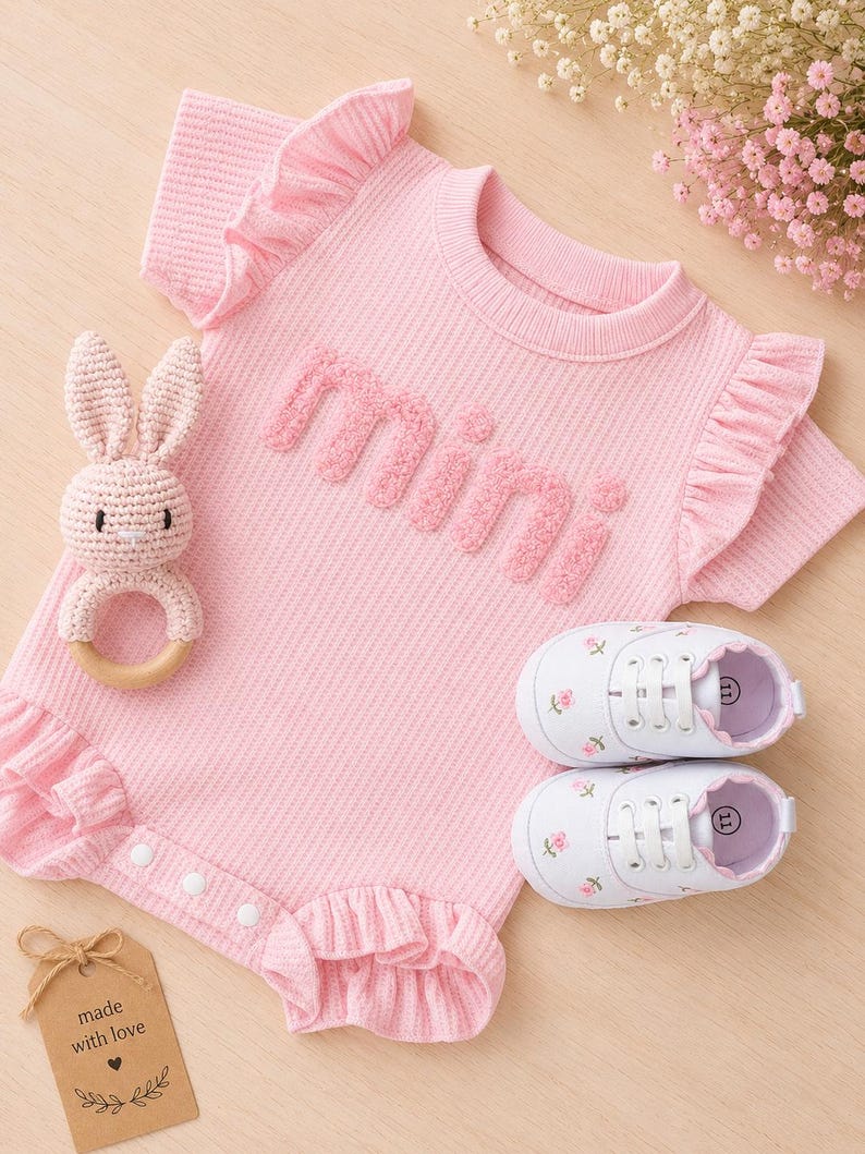 K&ouml;nnte beinhalten: Ein rosa Baby-Strampler mit R&uuml;schen&auml;rmeln und dem Wort "mini" in strukturierten Buchstaben. Enth&auml;lt eine H&auml;schen-Rassel, wei&szlig;e Babyschuhe mit Blumenstickerei und ein Etikett mit der Aufschrift "made with love".