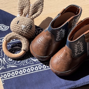 May include: A pair of brown and navy blue boots with white stitching details alongside a beige crocheted rabbit rattle featuring a smooth wooden ring, all resting on a dark blue bandana with a white paisley pattern.