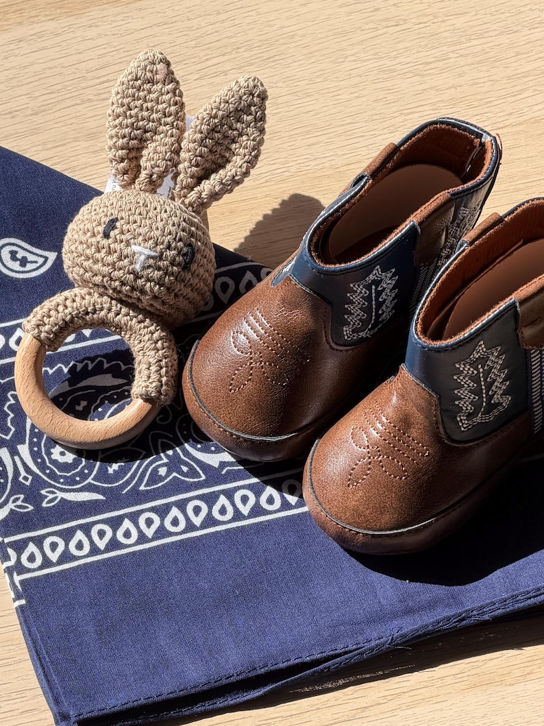 May include: A pair of brown and navy blue boots with white stitching details alongside a beige crocheted rabbit rattle featuring a smooth wooden ring, all resting on a dark blue bandana with a white paisley pattern.