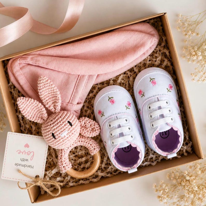 Peut inclure: Un coffret cadeau pour b&eacute;b&eacute; dans une bo&icirc;te en carton. L'ensemble comprend un chapeau oreilles de lapin rose, des chaussures de b&eacute;b&eacute; blanches avec broderie florale rose et un hochet lapin rose crochet&eacute; avec un anneau en bois. Une &eacute;tiquette indique "Handmade with love."