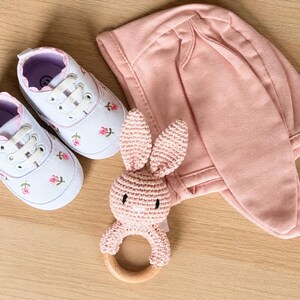 Peut inclure: Un ensemble cadeau pour b&eacute;b&eacute; comprenant des chaussures en toile blanche avec broderie florale rose, un bonnet &agrave; oreilles de lapin rose et un hochet lapin crochet&eacute; rose avec un anneau en bois. Sur une surface en bois clair.