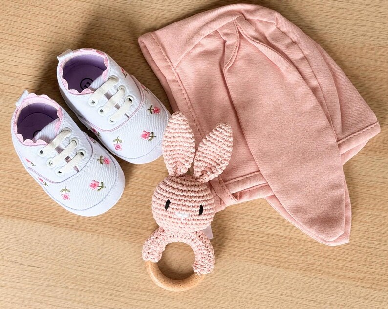 Peut inclure: Un ensemble cadeau pour b&eacute;b&eacute; comprenant des chaussures en toile blanche avec broderie florale rose, un bonnet &agrave; oreilles de lapin rose et un hochet lapin crochet&eacute; rose avec un anneau en bois. Sur une surface en bois clair.