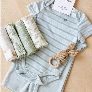 May include: A baby clothing set by Calvin Klein, featuring a light blue striped onesie and shorts, with a matching set of four muslin swaddles in shades of green and white. A crochet bunny rattle with a wooden ring is also included. The tag reads "Calvin Klein 3-Piece Set 6-9M". May include: A selection of baby essentials: a white Calvin Klein onesie, a crochet bunny rattle, brown baby shoes, and rolled muslin cloths. branded baby boy gift set. baby boy hamper set. baby boy Calvin Klein set.