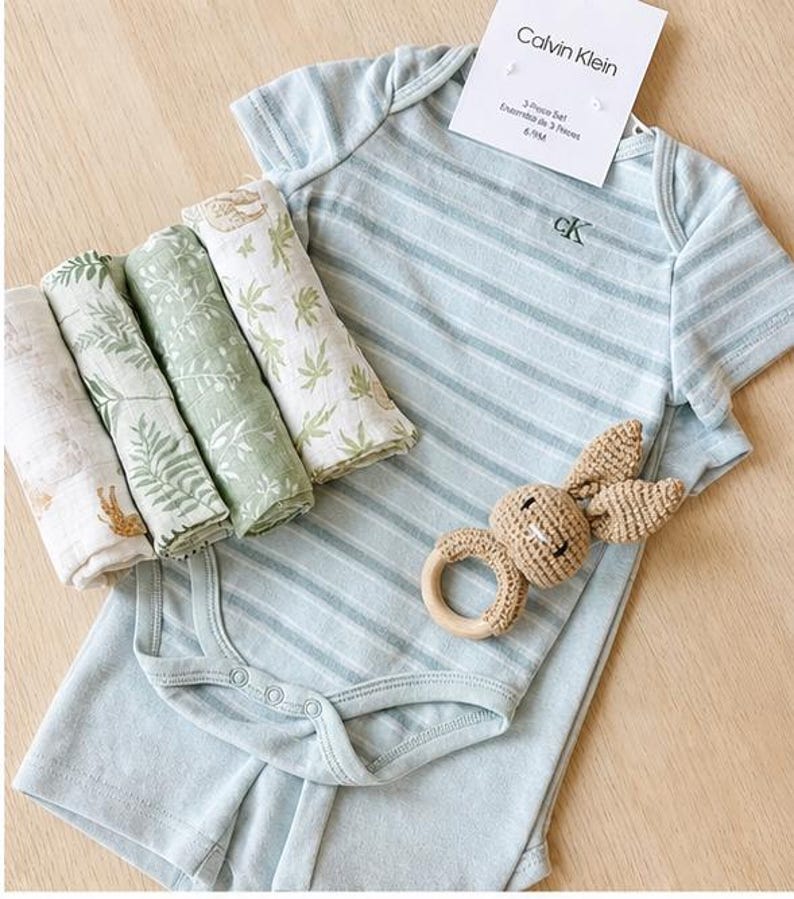 May include: A baby clothing set by Calvin Klein, featuring a light blue striped onesie and shorts, with a matching set of four muslin swaddles in shades of green and white. A crochet bunny rattle with a wooden ring is also included. The tag reads "Calvin Klein 3-Piece Set 6-9M". May include: A selection of baby essentials: a white Calvin Klein onesie, a crochet bunny rattle, brown baby shoes, and rolled muslin cloths. branded baby boy gift set. baby boy hamper set. baby boy Calvin Klein set.
