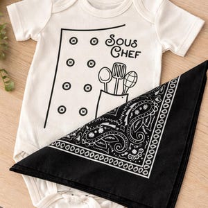Puede incluir: Un body blanco para beb&eacute; con un dise&ntilde;o de chaqueta de chef negra y la inscripci&oacute;n "Sous Chef". Un pa&ntilde;uelo negro con estampado de cachemira est&aacute; doblado junto al body. Se ilustran utensilios de cocina.