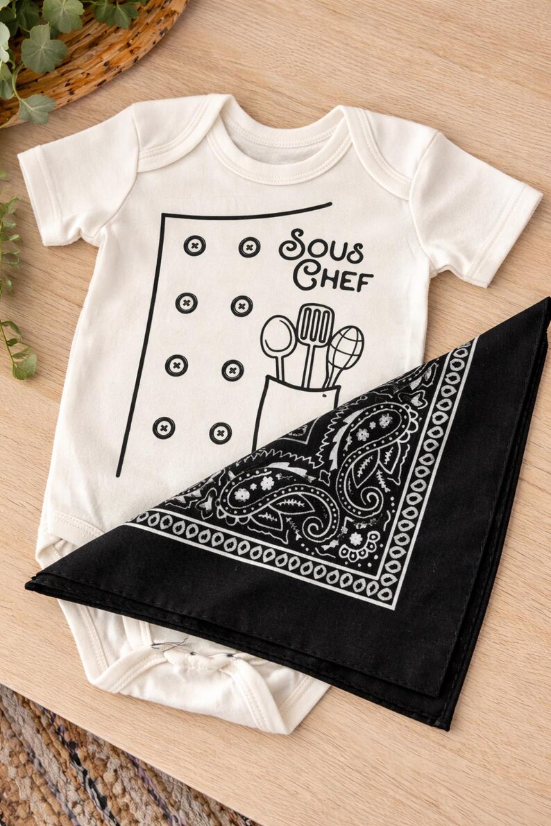 Puede incluir: Un body blanco para beb&eacute; con un dise&ntilde;o de chaqueta de chef negra y la inscripci&oacute;n "Sous Chef". Un pa&ntilde;uelo negro con estampado de cachemira est&aacute; doblado junto al body. Se ilustran utensilios de cocina.
