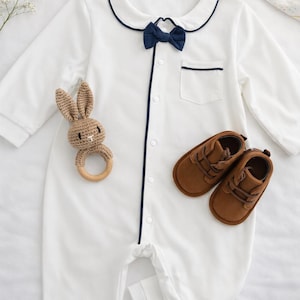 baby gentleman gift set bow onesie. White baby romper with navy blue trim and bow tie. Includes a small pocket and button-up front. A brown crochet bunny teether and brown leather baby shoes are also pictured. baby shower baby boy gift ideas.