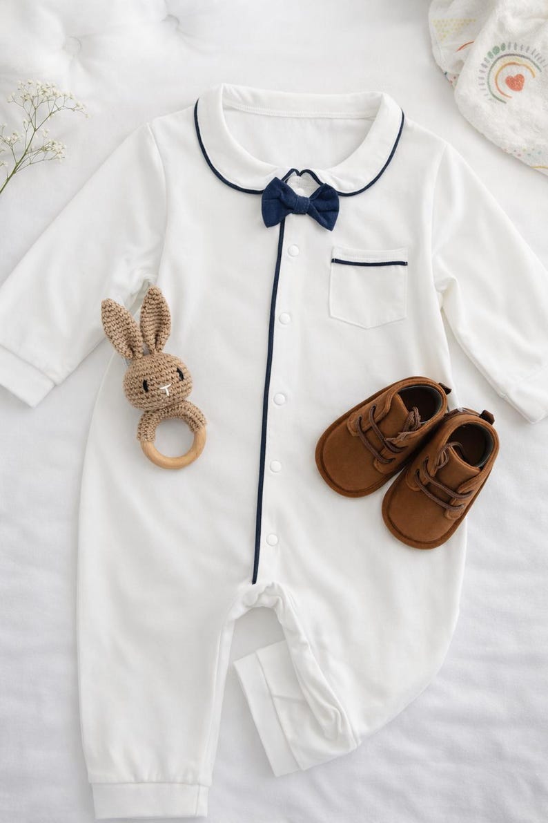 baby gentleman gift set bow onesie. White baby romper with navy blue trim and bow tie. Includes a small pocket and button-up front. A brown crochet bunny teether and brown leather baby shoes are also pictured. baby shower baby boy gift ideas.