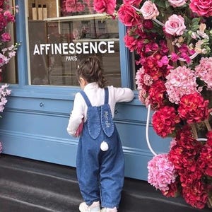 A blue denim overall with bunny ear detailing. The ears are embroidered with the name "Marielle" in white. A fluffy white pom-pom is attached to the back, resembling a bunny tail. The overall is designed for a child. Cute bunny Easter overall