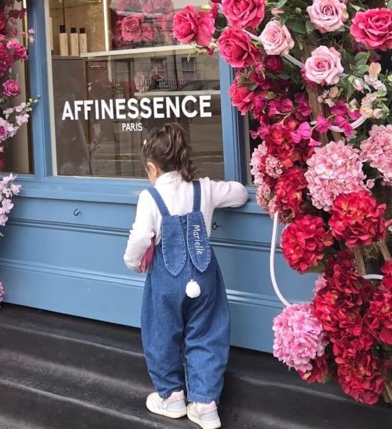 A blue denim overall with bunny ear detailing. The ears are embroidered with the name "Marielle" in white. A fluffy white pom-pom is attached to the back, resembling a bunny tail. The overall is designed for a child. Cute bunny Easter overall