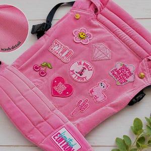 K&ouml;nnte beinhalten: Rosa Babytrage mit mehreren gestickten Aufn&auml;hern, darunter eine Blume und ein Herz. Die Trage hat schwarze Gurte und Schnallen, mit dem Text "Limited Edition". Der Stoff hat ein Wei&szlig;waschdetail.