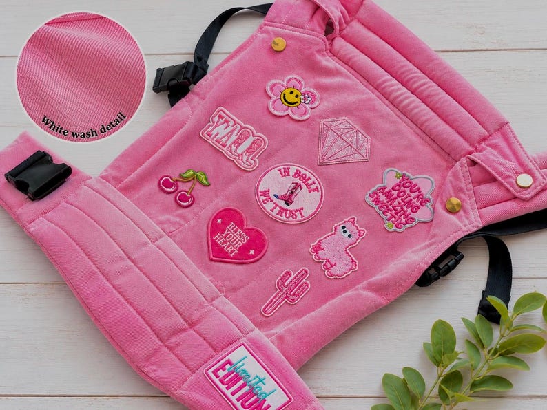 K&ouml;nnte beinhalten: Rosa Babytrage mit mehreren gestickten Aufn&auml;hern, darunter eine Blume und ein Herz. Die Trage hat schwarze Gurte und Schnallen, mit dem Text "Limited Edition". Der Stoff hat ein Wei&szlig;waschdetail.