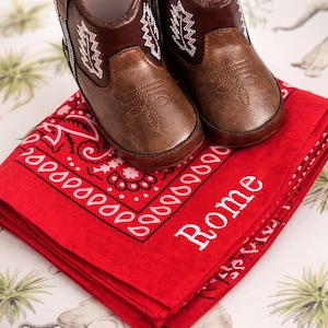 Pode incluir: Um par de botinhas de cowboy castanhas de couro para beb&eacute;, com detalhes de costura brancos, pousadas numa bandana vermelha dobrada com o nome "Rome" em branco. A bandana tem uma borda paisley.