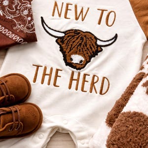 Peut inclure: Ensemble de v&ecirc;tements pour b&eacute;b&eacute; blanc et marron avec un motif de vache des Highlands. L'ensemble comprend une barboteuse avec le texte "NEW TO THE HERD", des chaussures marron, un bandana et une couverture &agrave; imprim&eacute; vache.