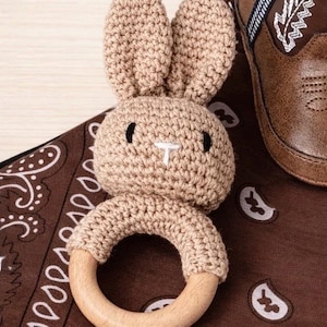 May include: A tan crochet bunny-shaped baby rattle with a wooden ring. The bunny has black eyes and a white nose. The rattle is on a brown paisley patterned cloth, next to a brown leather baby boot.