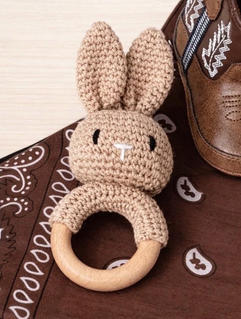 May include: A tan crochet bunny-shaped baby rattle with a wooden ring. The bunny has black eyes and a white nose. The rattle is on a brown paisley patterned cloth, next to a brown leather baby boot.