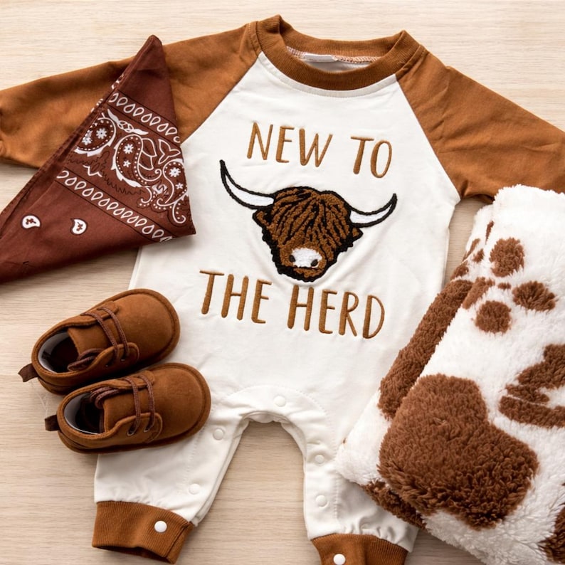 Peut inclure: Ensemble de v&ecirc;tements pour b&eacute;b&eacute; comprenant un body blanc et marron avec l'inscription "NEW TO THE HERD" et un motif de vache des Highlands. Comprend un bandana marron, des chaussures de b&eacute;b&eacute; marron et une couverture &agrave; imprim&eacute; vache.