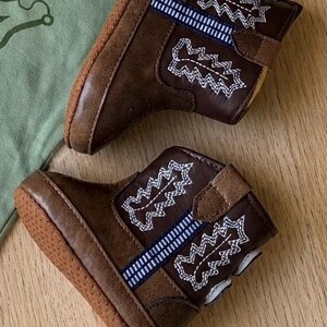 May include: A pair of brown baby cowboy boots with white and blue embroidered details. The boots are made of a leather-like material and have a rubber sole. A green t-shirt with the text "DY'S" is in the background.