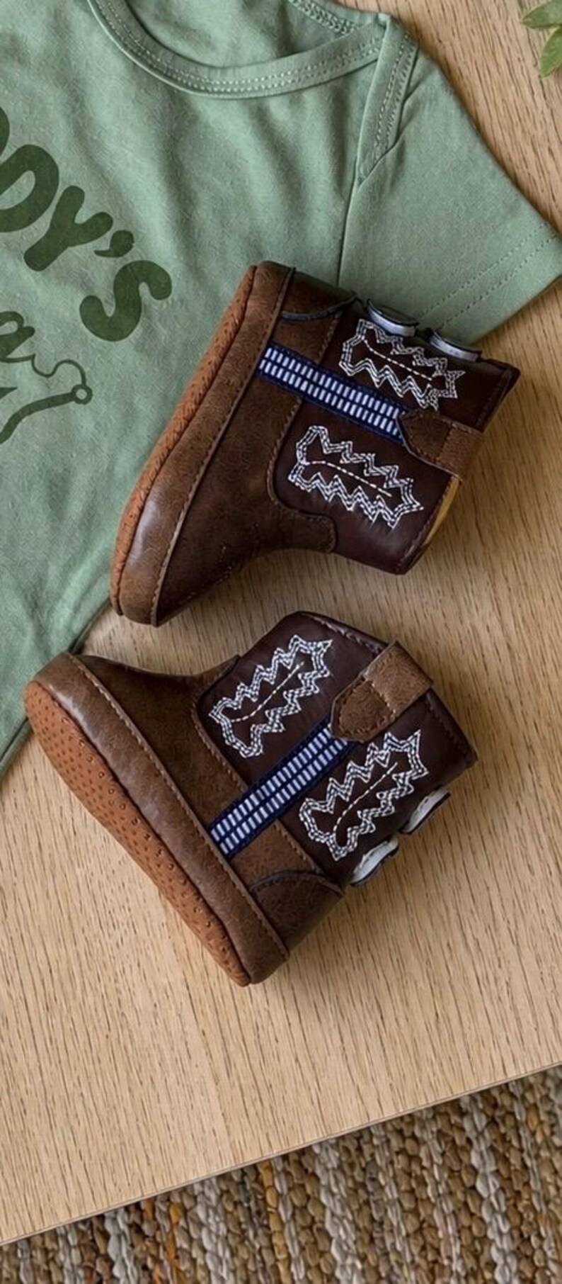 May include: A pair of brown baby cowboy boots with white and blue embroidered details. The boots are made of a leather-like material and have a rubber sole. A green t-shirt with the text "DY'S" is in the background.