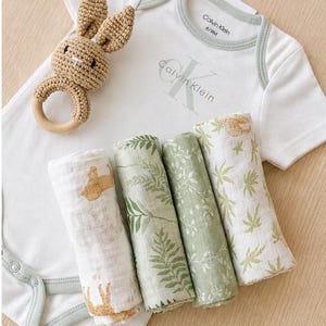 May include: A baby gift set featuring a white Calvin Klein onesie, four rolled muslin swaddles with botanical and animal prints, and a crochet bunny rattle. The onesie is size 6/9M.