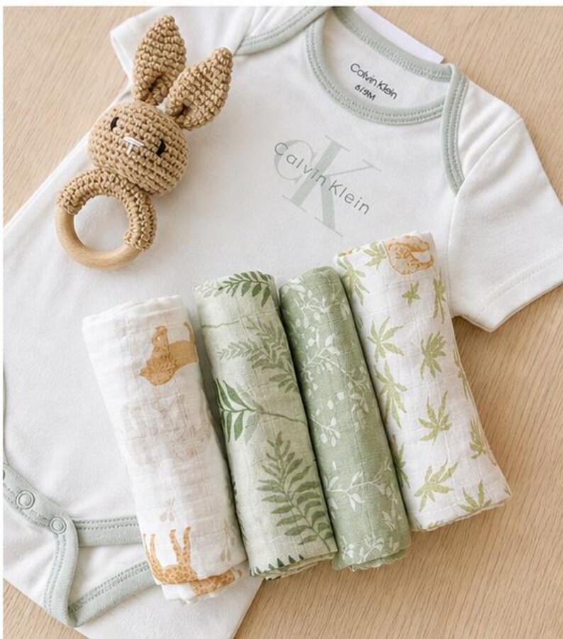 May include: A baby gift set featuring a white Calvin Klein onesie, four rolled muslin swaddles with botanical and animal prints, and a crochet bunny rattle. The onesie is size 6/9M.