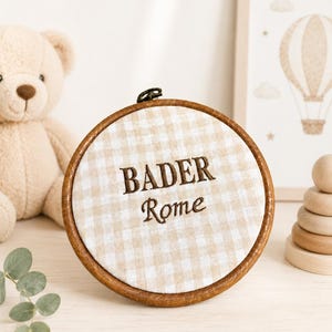 May include: A round, wooden embroidery hoop with the name "BADER" and the word "Rome" embroidered in brown thread on a beige and white checkered fabric. A teddy bear and a framed hot air balloon art piece are in the background.