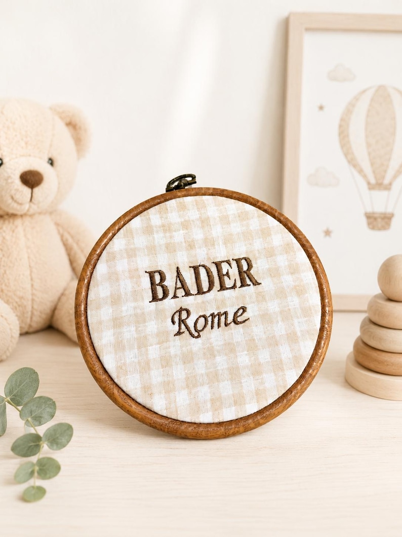 May include: A round, wooden embroidery hoop with the name "BADER" and the word "Rome" embroidered in brown thread on a beige and white checkered fabric. A teddy bear and a framed hot air balloon art piece are in the background.