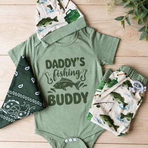 May include: A baby outfit set in shades of green and cream, featuring a short-sleeved bodysuit with the text "DADDY'S fishing BUDDY" and a fish graphic. The set includes a matching hat, bandana, and patterned pants with fishing-themed illustrations.
