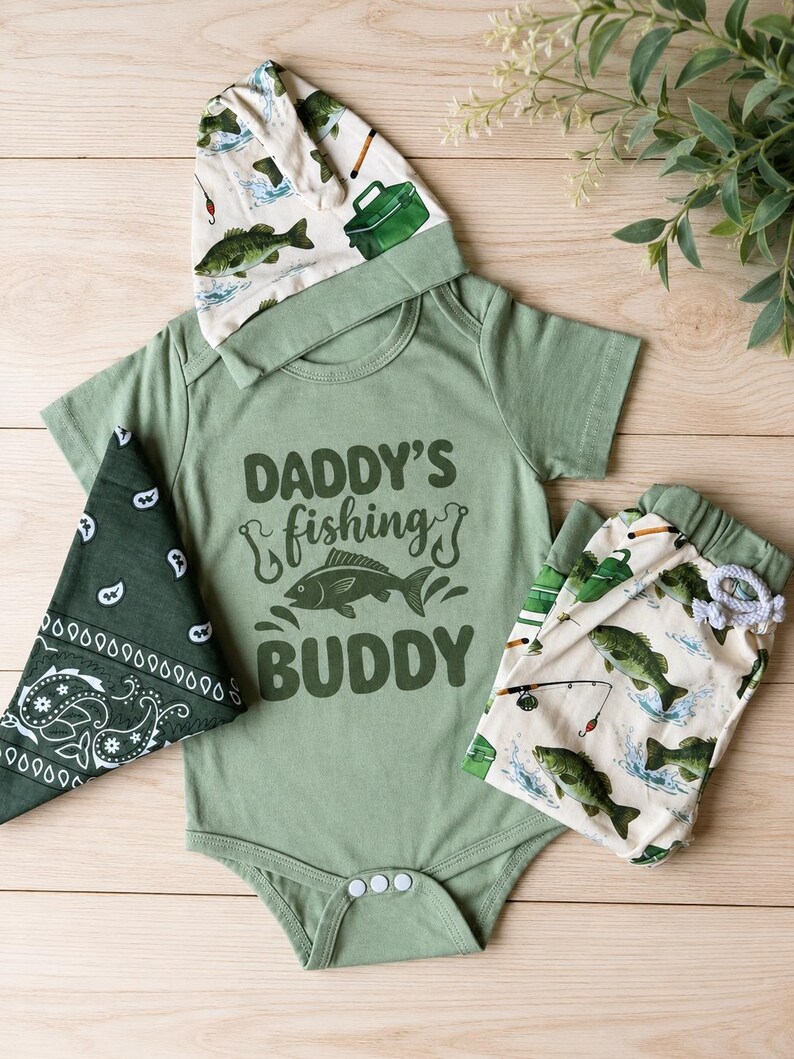 May include: A baby outfit set in shades of green and cream, featuring a short-sleeved bodysuit with the text "DADDY'S fishing BUDDY" and a fish graphic. The set includes a matching hat, bandana, and patterned pants with fishing-themed illustrations.