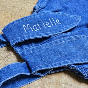 A blue denim overall with bunny ear detailing. The ears are embroidered with the name "Marielle" in white. A fluffy white pom-pom is attached to the back, resembling a bunny tail. The overall is designed for a child. Cute bunny Easter overall
