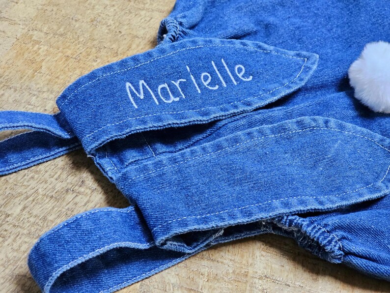 A blue denim overall with bunny ear detailing. The ears are embroidered with the name "Marielle" in white. A fluffy white pom-pom is attached to the back, resembling a bunny tail. The overall is designed for a child. Cute bunny Easter overall