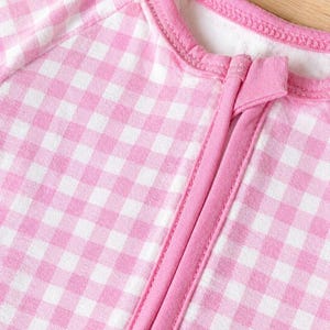 May include: Close-up of a pink and white gingham-patterned baby sleeper. The sleeper has a pink zipper and trim, and a round neckline. The fabric is soft and appears to be cotton or a similar material.