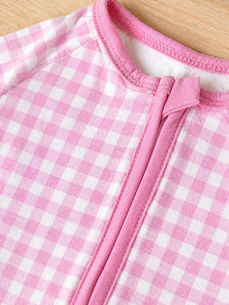 May include: Close-up of a pink and white gingham-patterned baby sleeper. The sleeper has a pink zipper and trim, and a round neckline. The fabric is soft and appears to be cotton or a similar material.