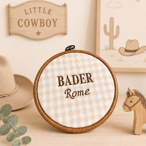 May include: A round embroidery hoop with a tan and white gingham fabric. The text "BADER Rome" is embroidered in brown thread. A wooden horse figurine and a "Little Cowboy" sign are also visible.