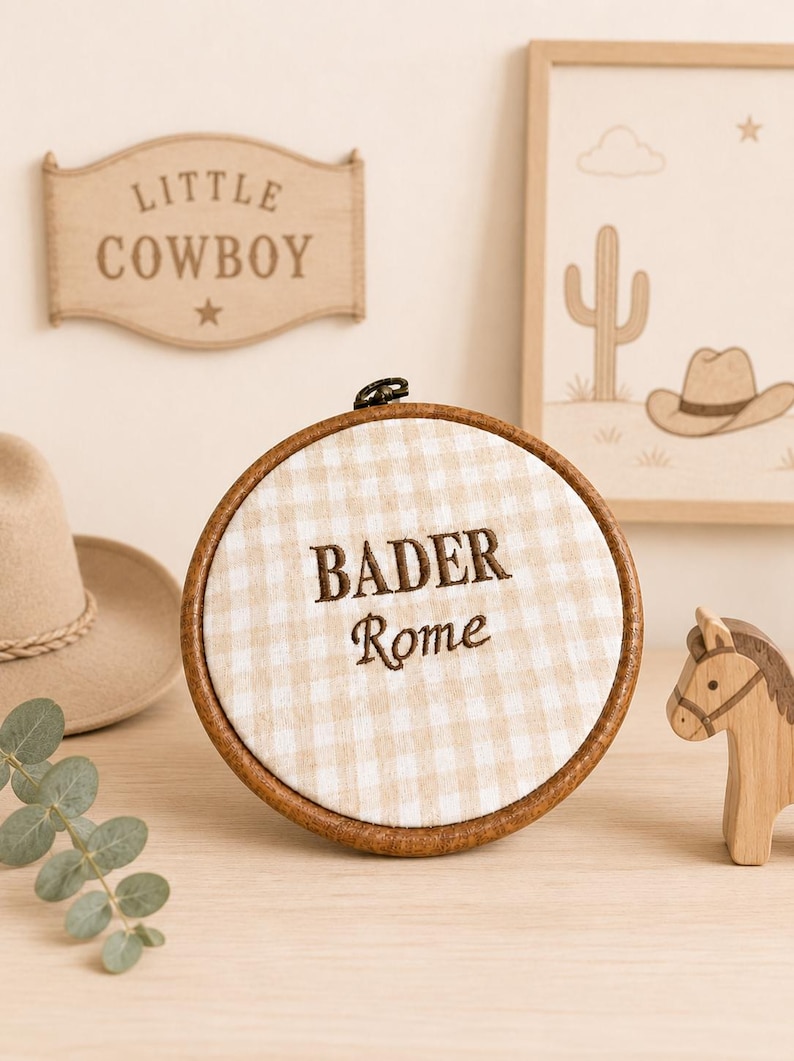 May include: A round embroidery hoop with a tan and white gingham fabric. The text "BADER Rome" is embroidered in brown thread. A wooden horse figurine and a "Little Cowboy" sign are also visible.