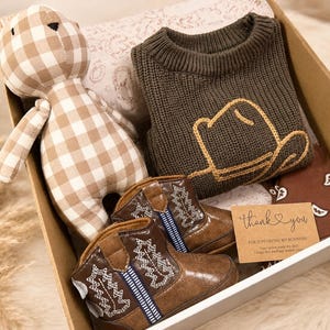 May include: A gift box containing a beige and white gingham teddy bear, a dark olive green knit sweater with a yellow embroidered cowboy hat outline, brown faux leather cowboy boots with white stitching, and a thank you card with text.